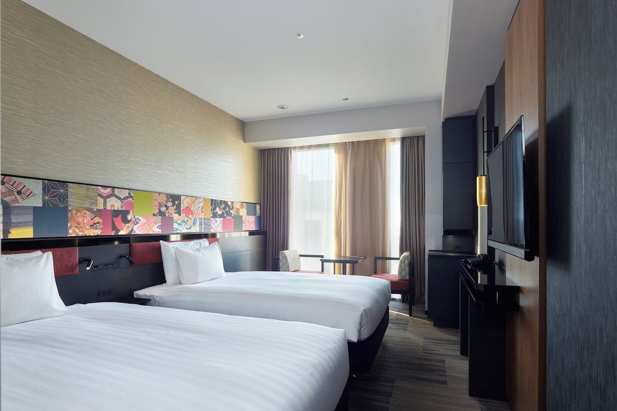 A hotel room with two beds, a TV, a desk, and a large window with curtains.