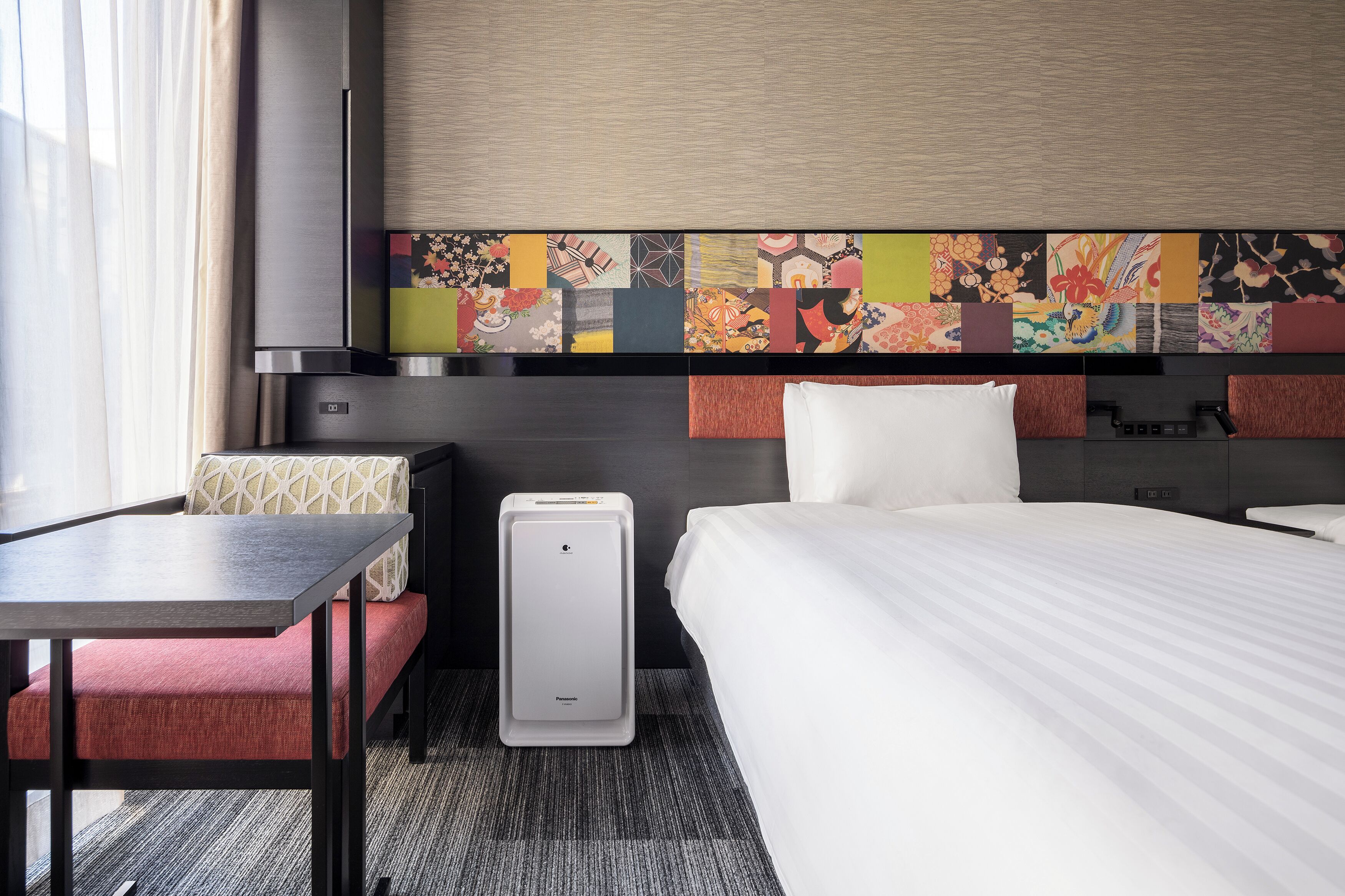 Una camera d'albergo con un letto, una scrivania, una sedia, una valigia e un quadro colorato appeso al muro.