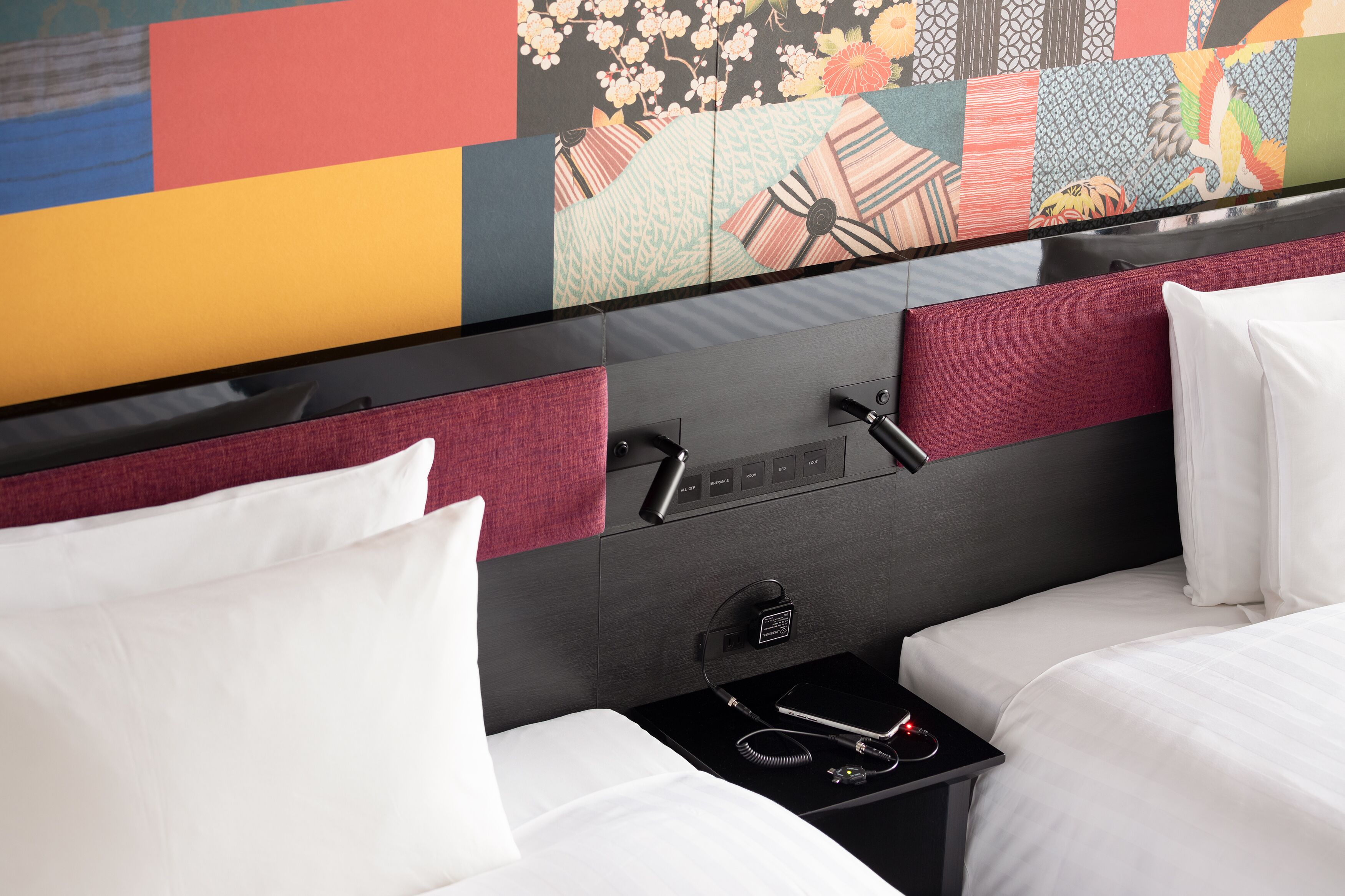 Ein Hotelzimmer mit einem großen Spiegel, zwei Betten mit weißer Bettwäsche, einem kleinen Nachttisch mit Telefon und Ladegerät sowie einem farbenfrohen Wandbild.