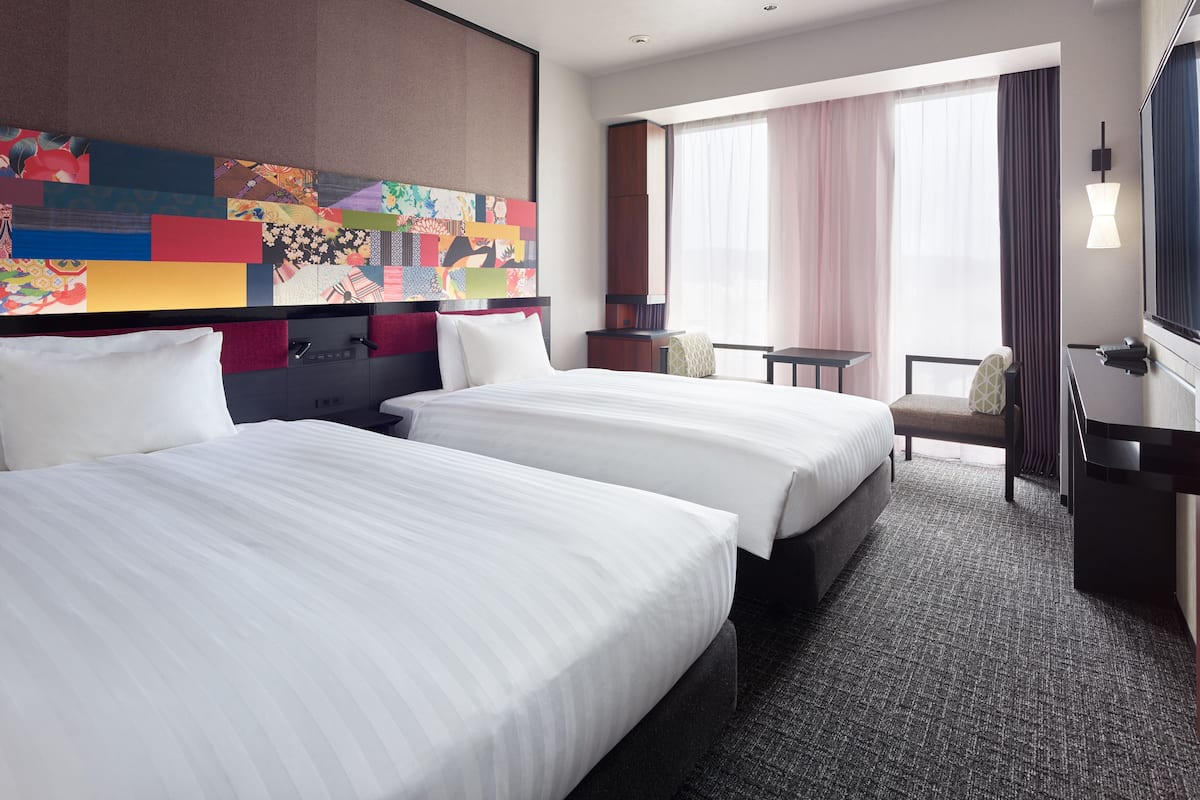 A hotel room with two beds, a large colorful artwork on the wall, a desk with a lamp, and a view of the outside through large windows.