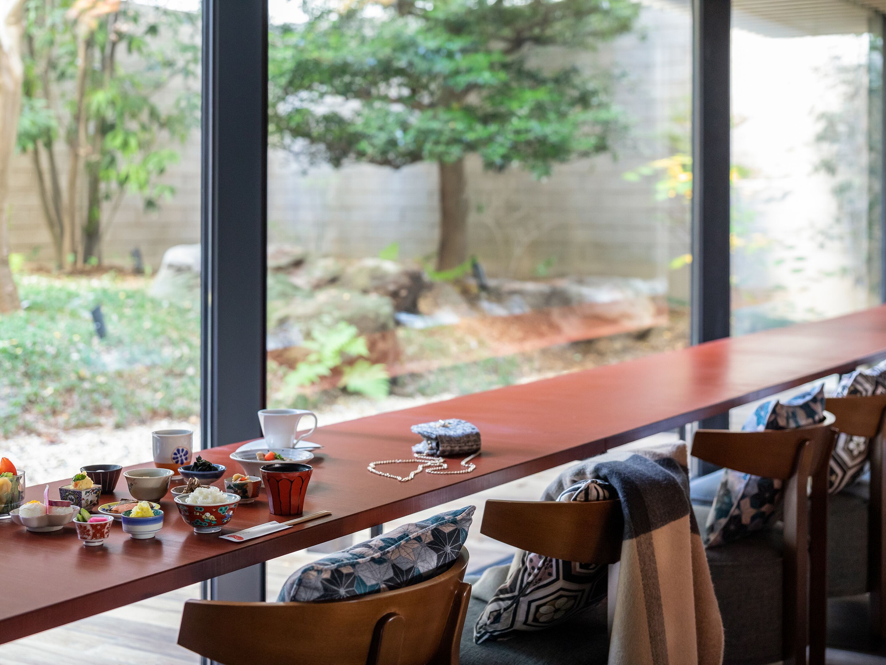A dining area with a long table, various dishes, and a view of a garden through large windows.