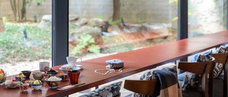 A dining area with a long table, various dishes, and a view of a garden through large windows.