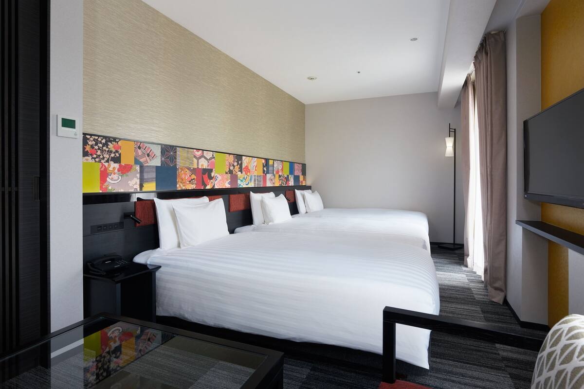 A hotel room with two beds, a desk, a TV, and a colorful abstract art piece on the wall.