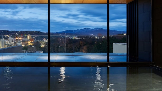 A modern interior with large windows offering a view of a cityscape and mountains at dusk. - Mitsui Garden Hotel Kanazawa (Kanazawa)