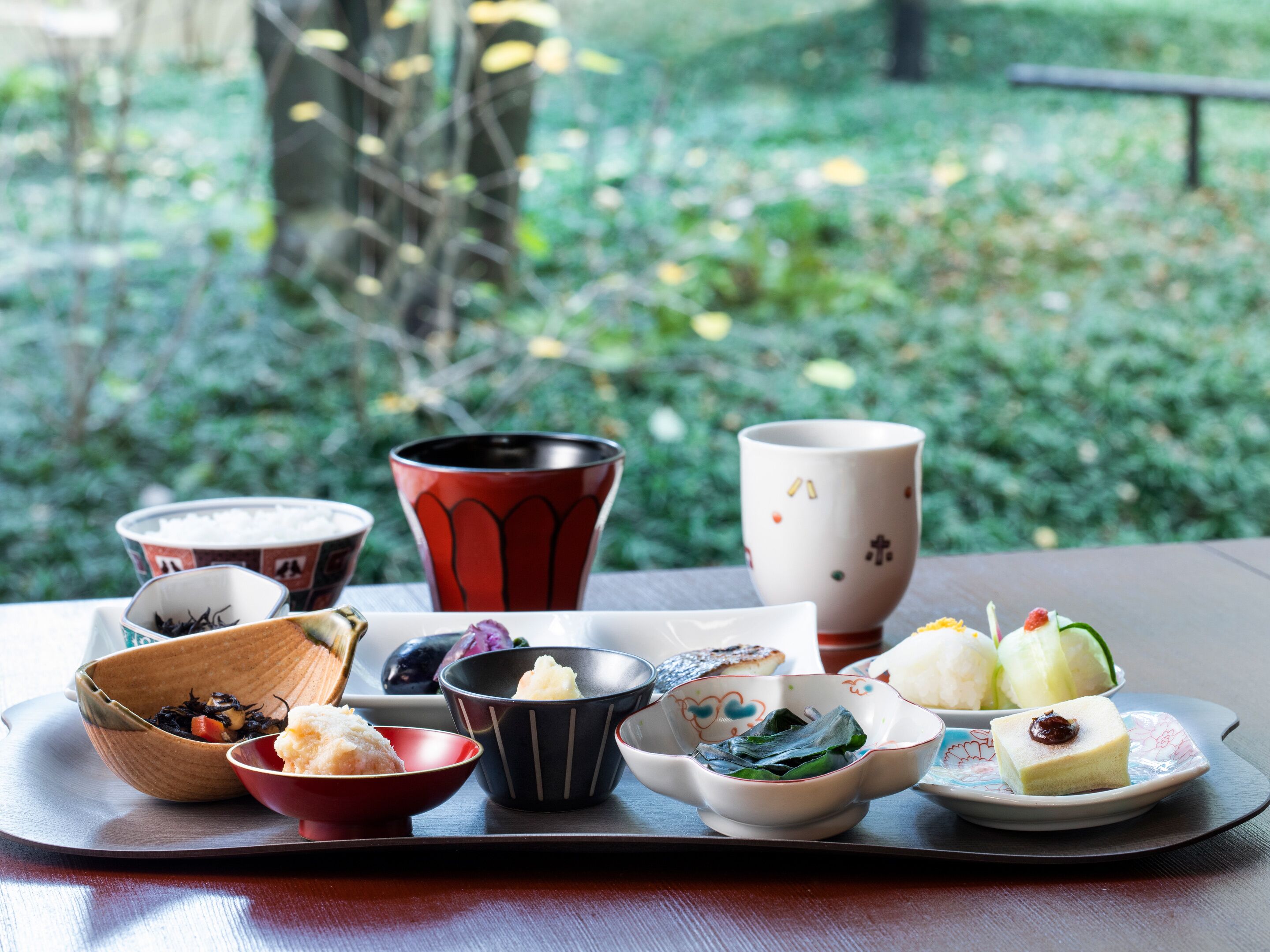 Tarjottimella on erilaisia japanilaisia ruokia, kuten sushia, riisiä ja marinoituja kasviksia, ja se on asetettu pöydälle, josta avautuu näkymä vehreään maisemaan.
