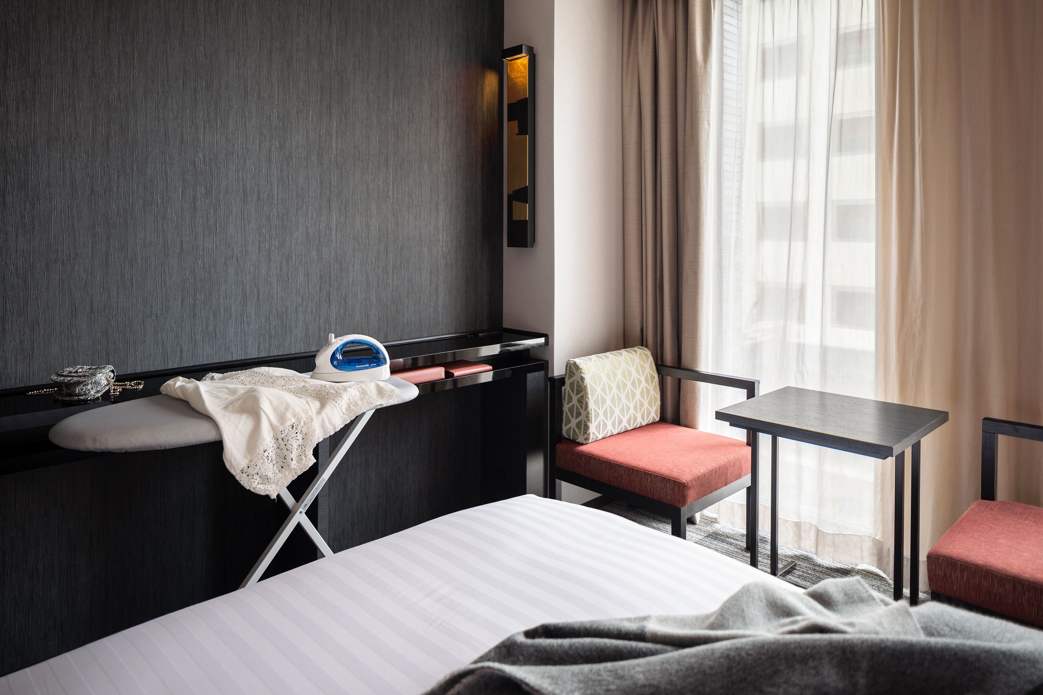 A hotel room with a bed, a small table, a chair, a desk with a lamp, and a window with curtains.