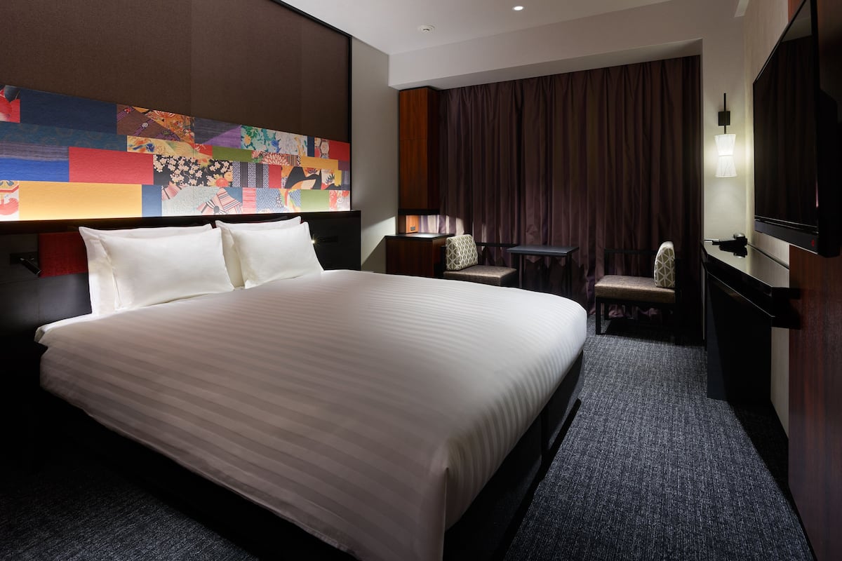 A hotel room with a large bed, a desk, a chair, a television, and a colorful artwork on the wall.