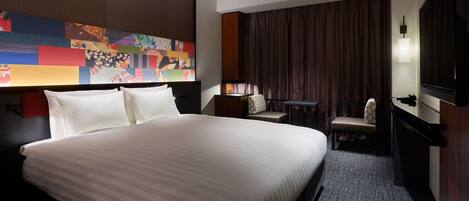 A hotel room with a large bed, a desk, a chair, a television, and a colorful artwork on the wall.