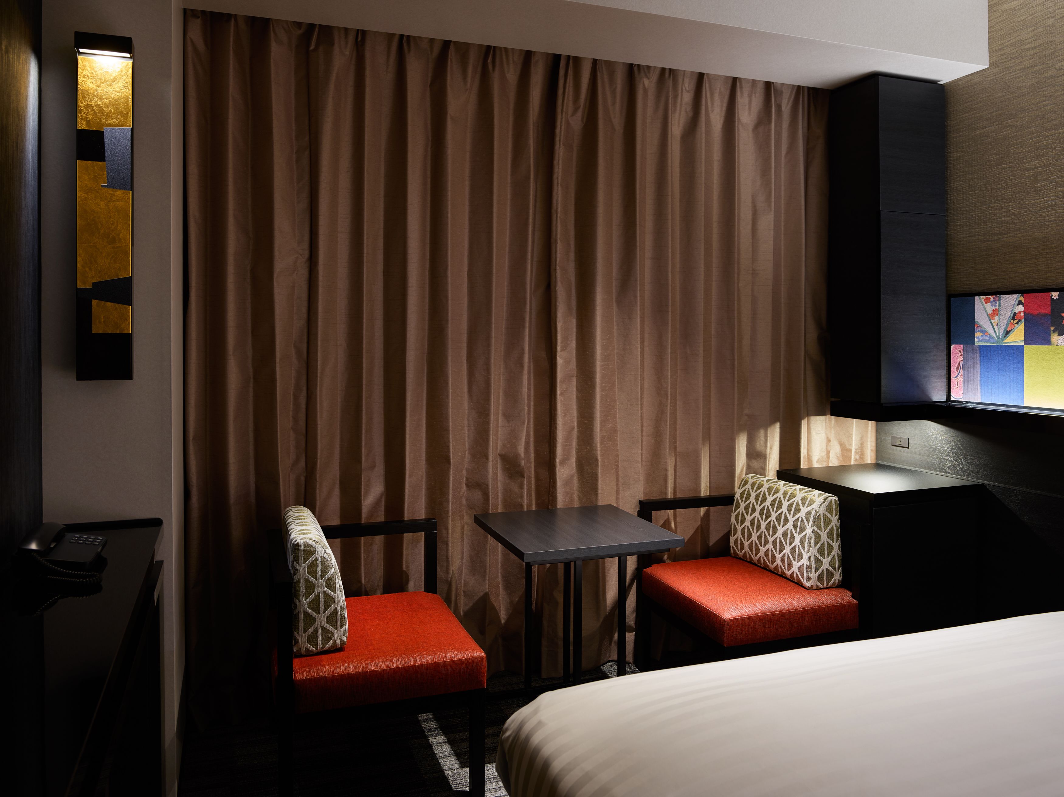 A hotel room with a bed, two red upholstered chairs, a small table, a TV, and a curtain.