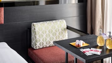 A hotel room with a bed, a sofa, a small table with toiletries, and a pillow with a geometric pattern.