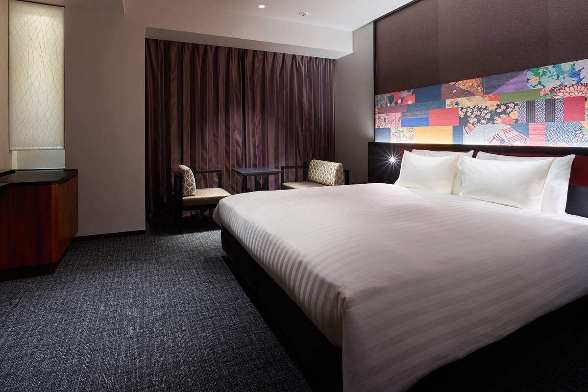 A hotel room with a large bed, a desk, a chair, and a colorful artwork on the wall.