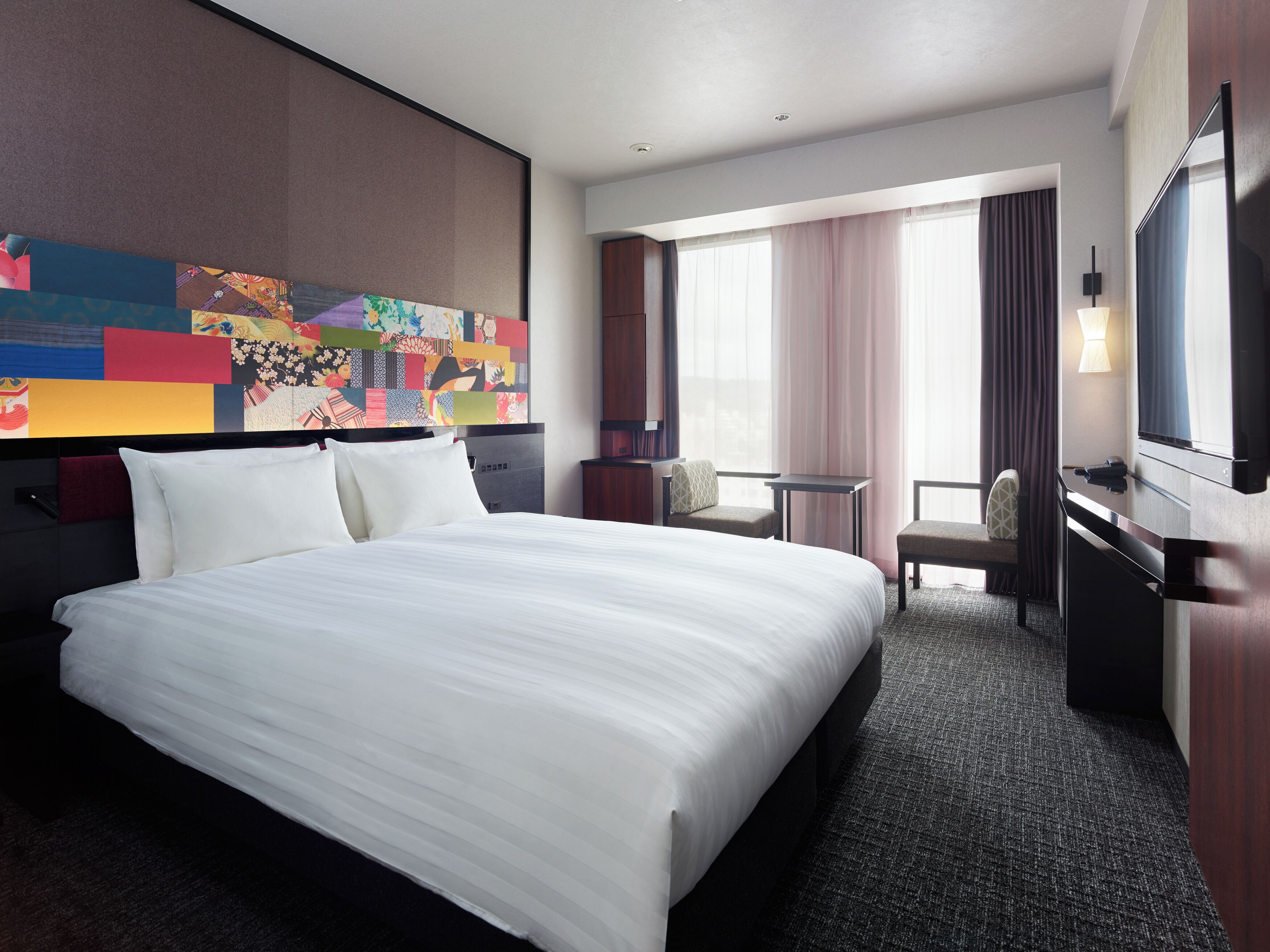A hotel room with a large bed, a desk, a chair, a television, and a colorful wall art piece.