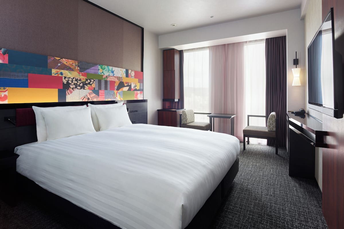 A hotel room with a large bed, a desk, a chair, a television, and a colorful wall art piece.