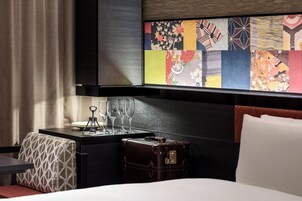 Habitación de hotel con cama, mesita de noche con vasos, maleta y un cuadro de colores en la pared.