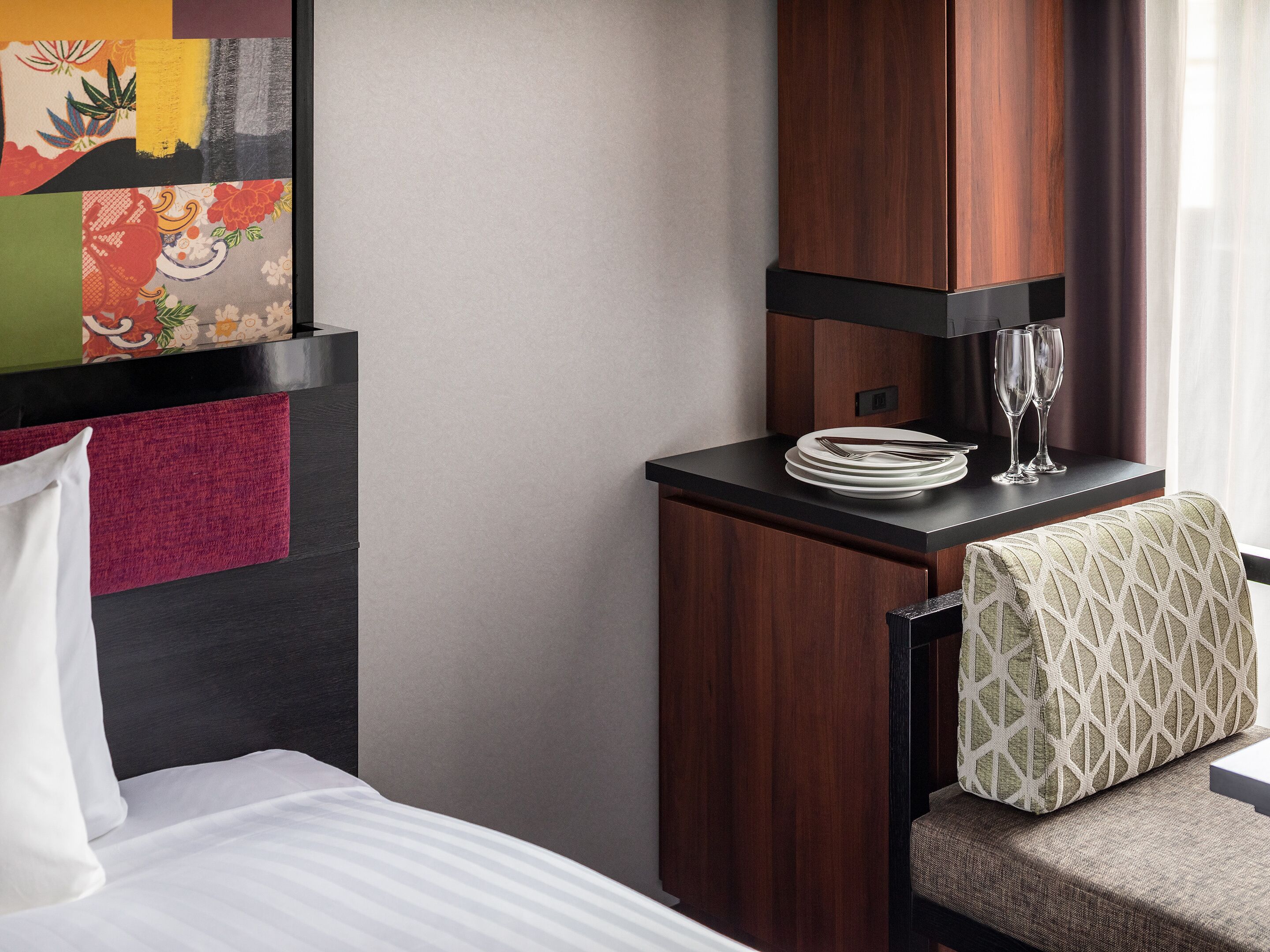 A hotel room with a bed, a small table with a plate and glasses, a chair with a patterned cushion, and a wall with a colorful artwork.