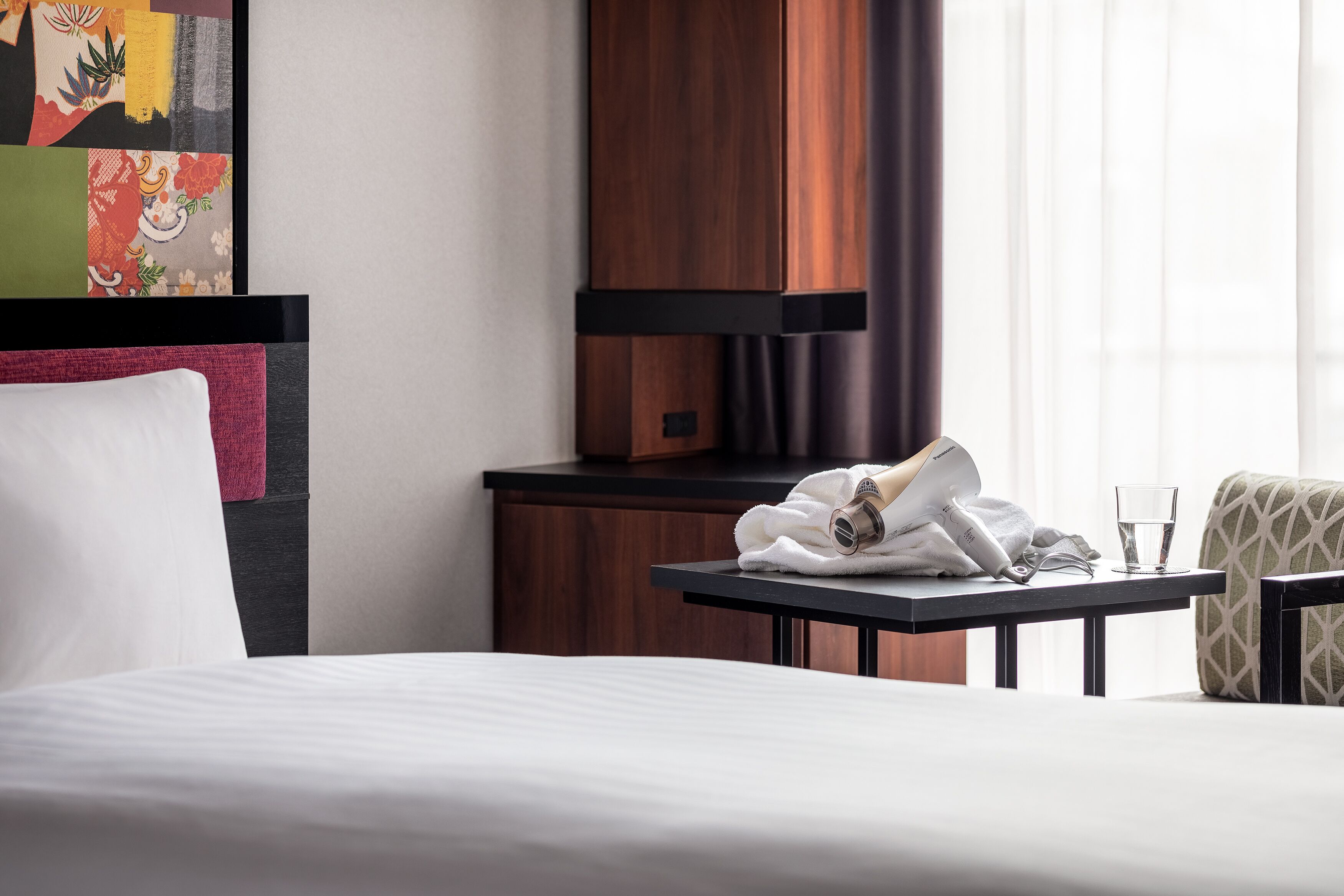 A hotel room with a neatly made bed, a small table with a towel and a glass of water, and a chair.