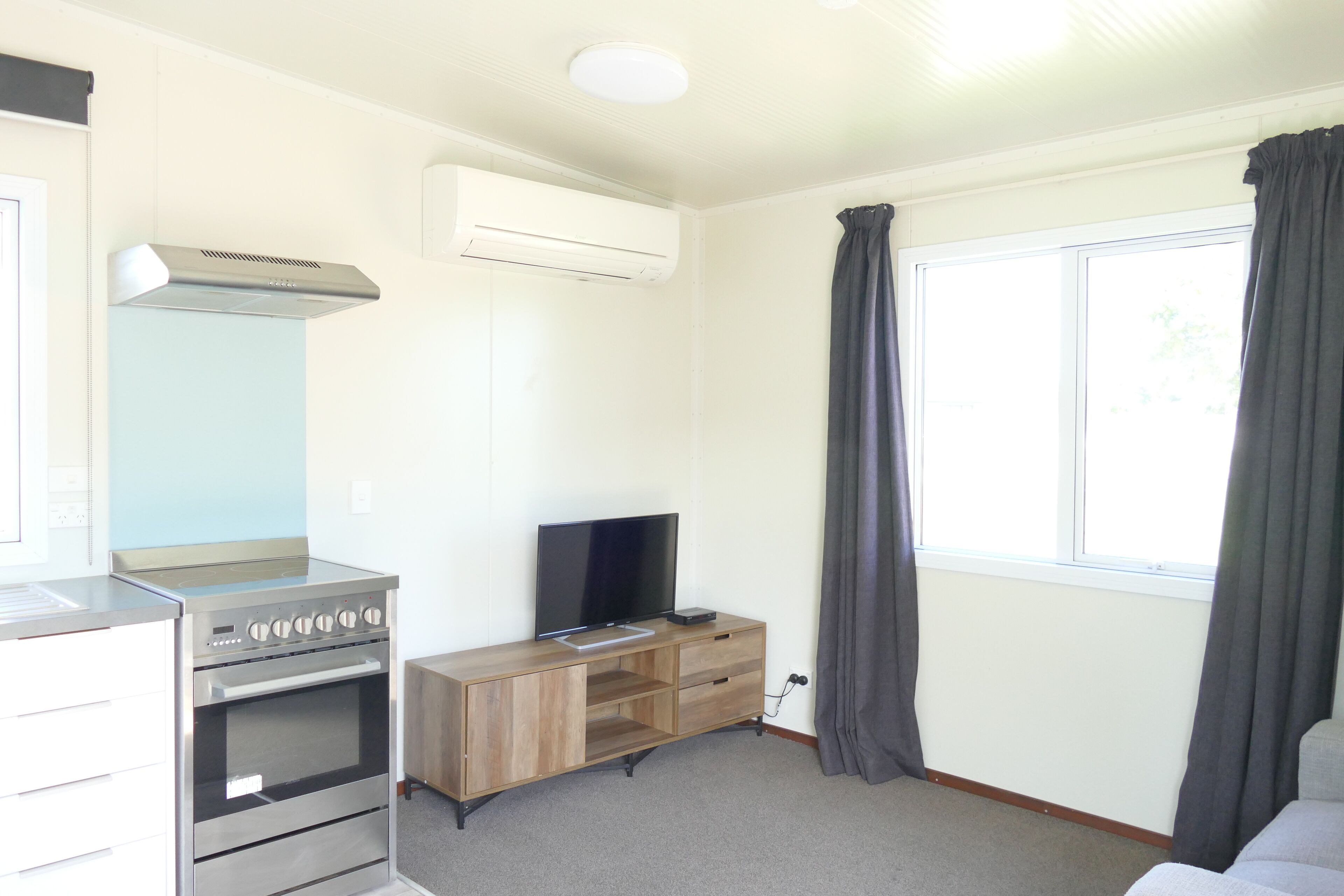 Un espacio habitable compacto con kitchenette, televisor de pantalla plana y un amplio ventanal con cortinas.
