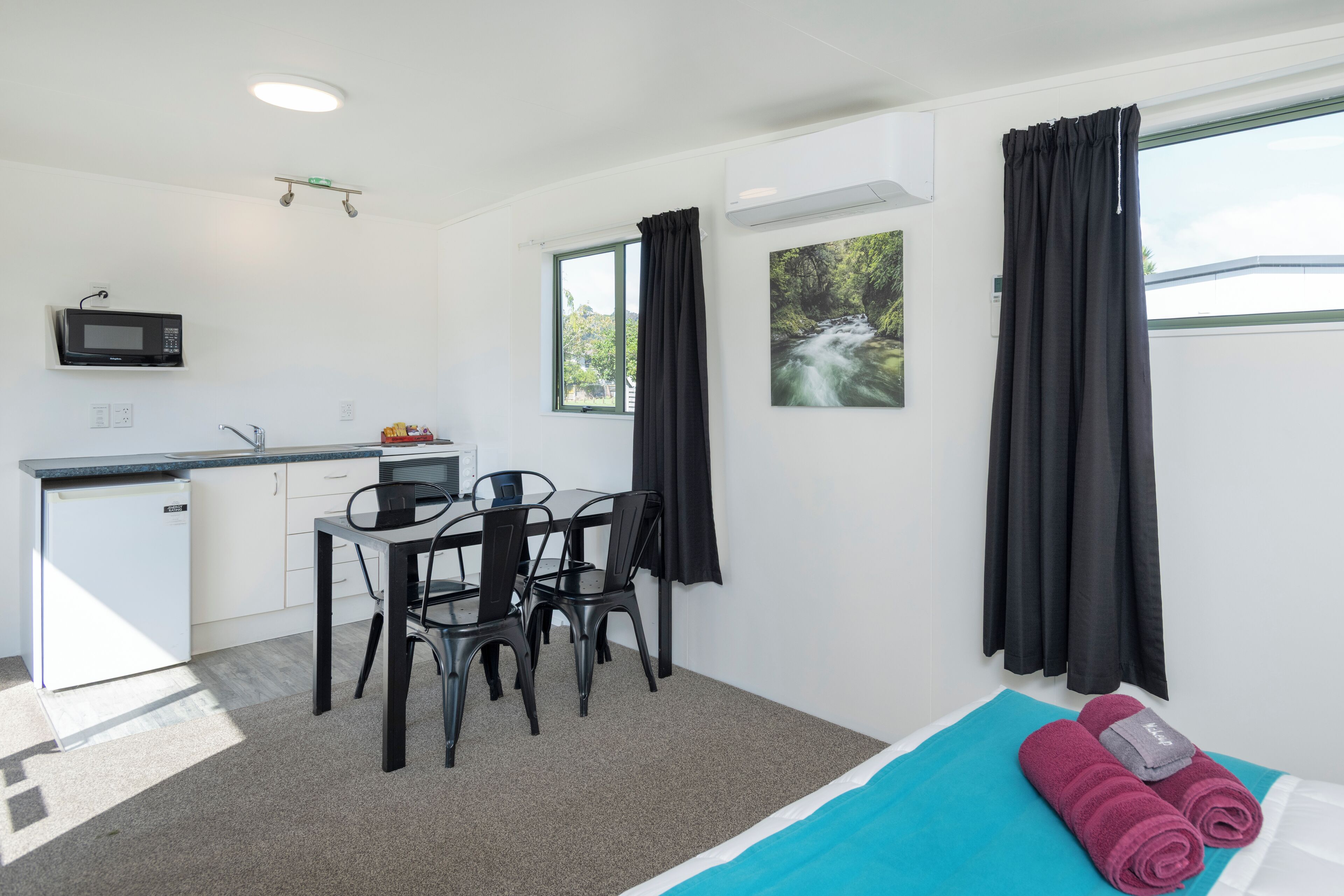 Ein gemütliches Zimmer mit Küchenzeile, einem Essbereich, einem Bett mit blau-weißer Bettdecke und einem Fenster mit Blick auf Grünflächen.