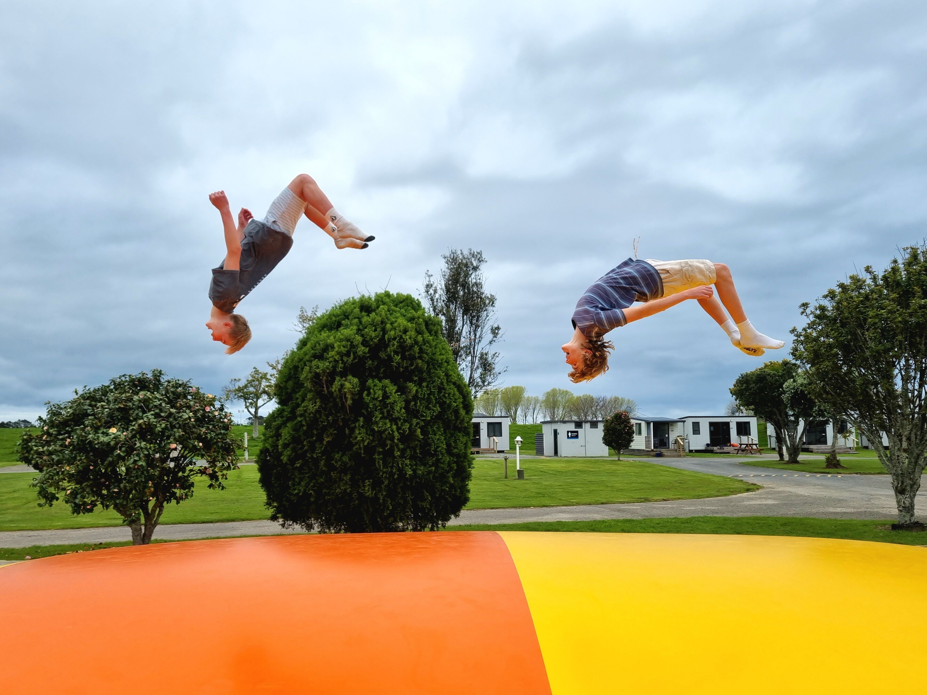 Two people jumping on a colorful playground with a grassy area and trees in the background.