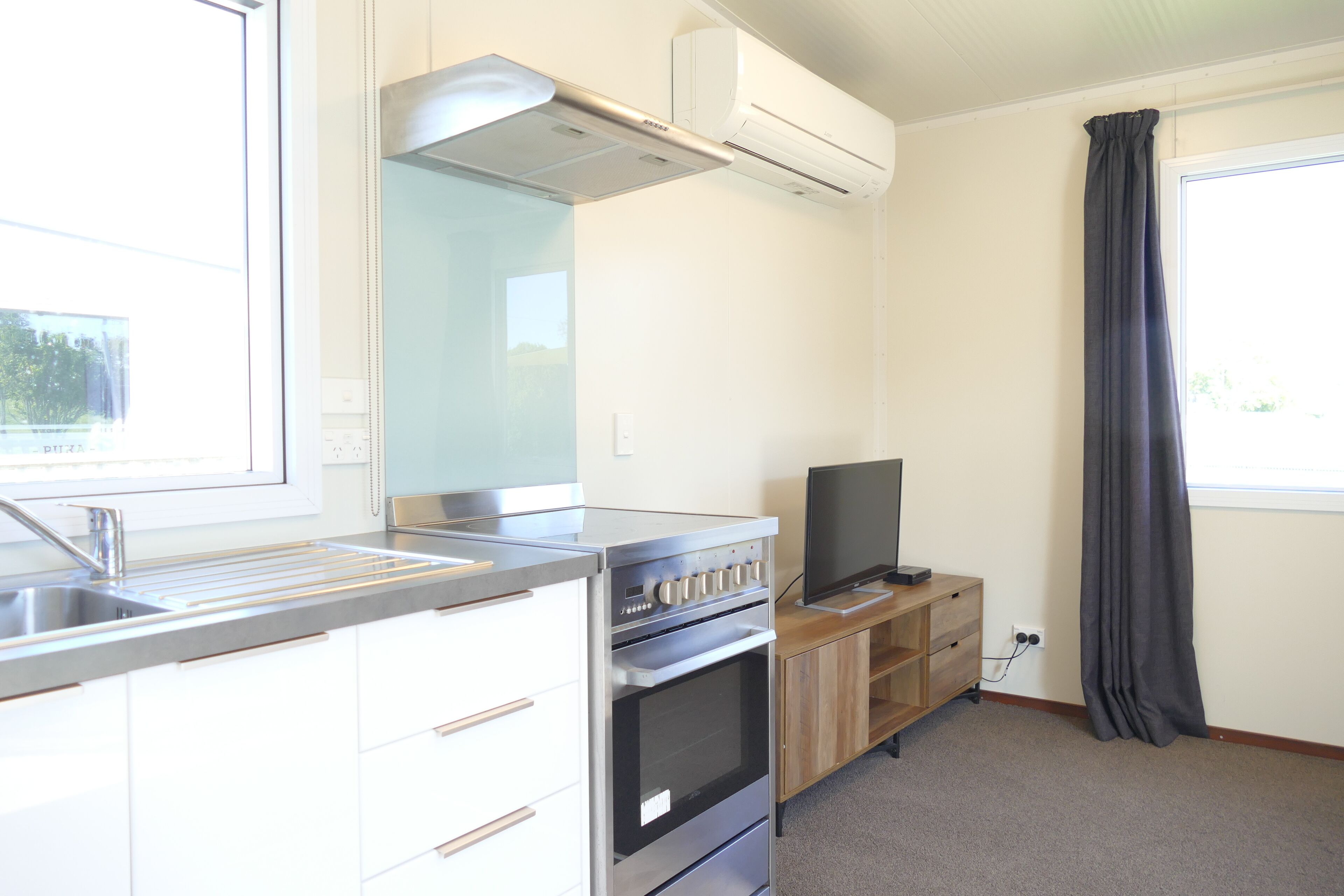 A compact kitchen with a built-in oven, a microwave, and a sink. There is a window with a curtain and a flat-screen TV on a wooden stand.