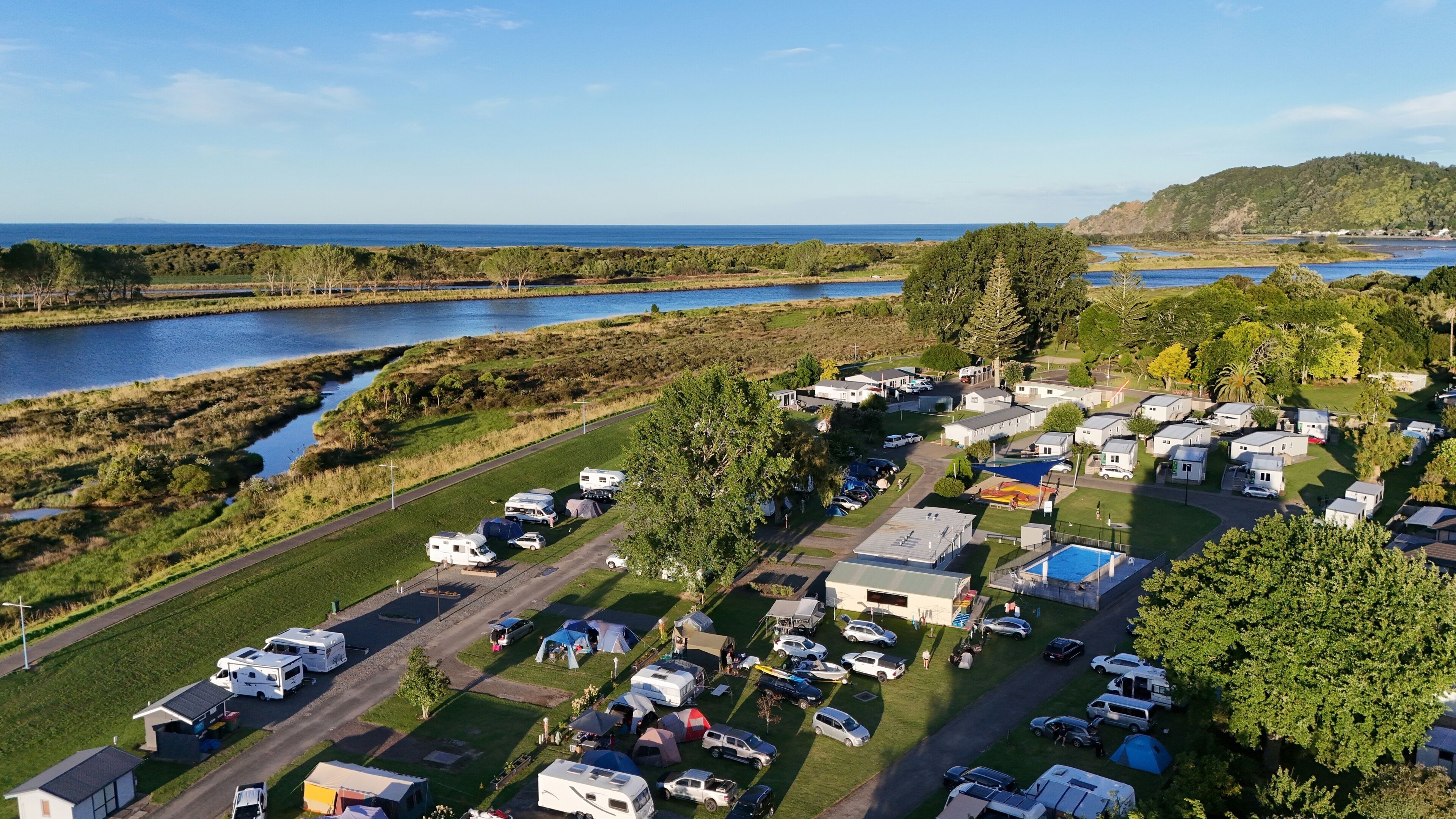 An aerial view of a campground with RVs and cars parked, a swimming pool, and a river in the background.