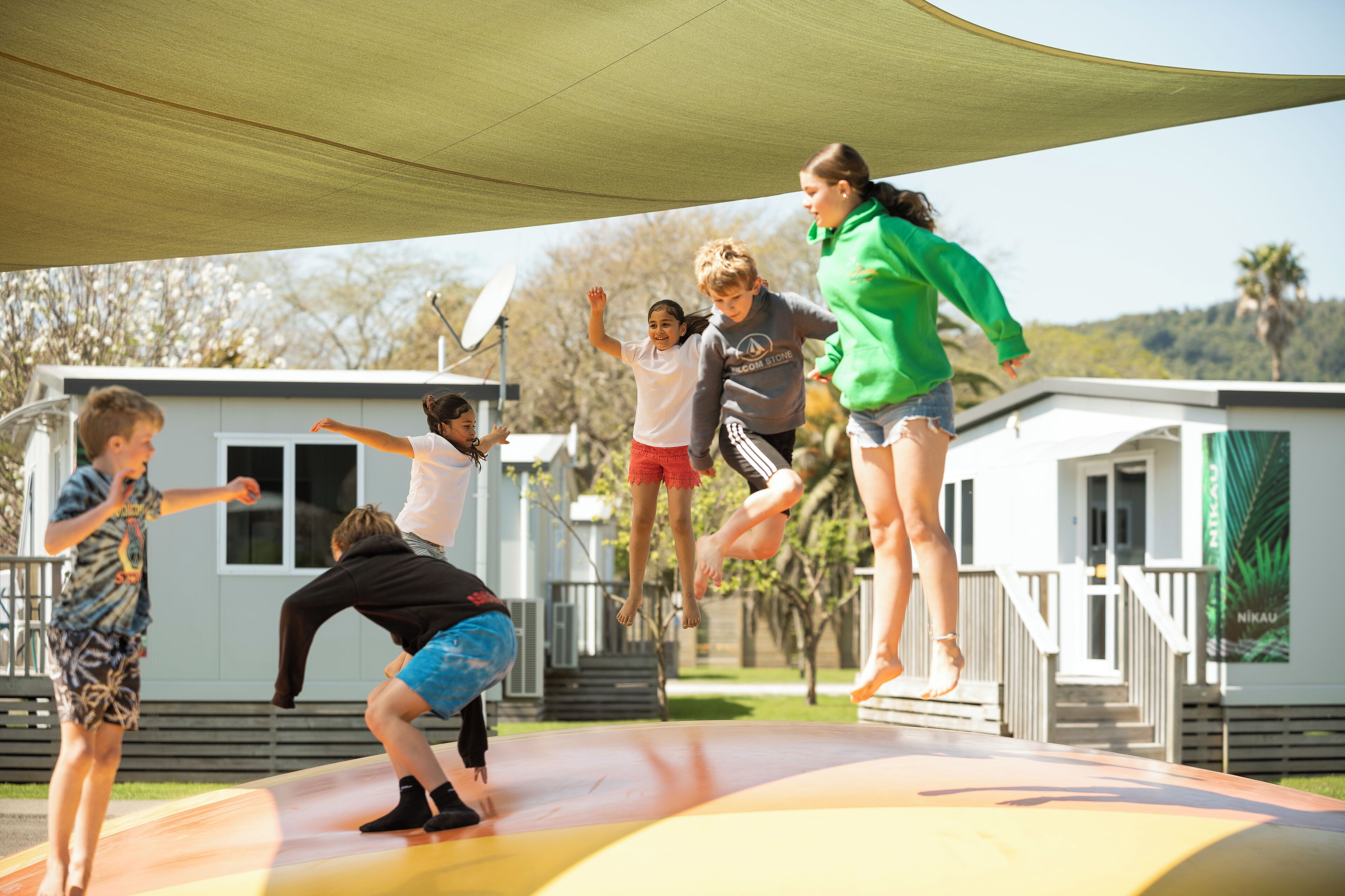 A group of children playing on a trampoline under a shaded area with small cabins in the background.