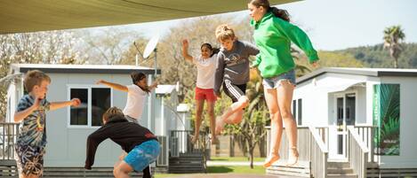 A group of children playing on a trampoline under a shaded area with small cabins in the background.