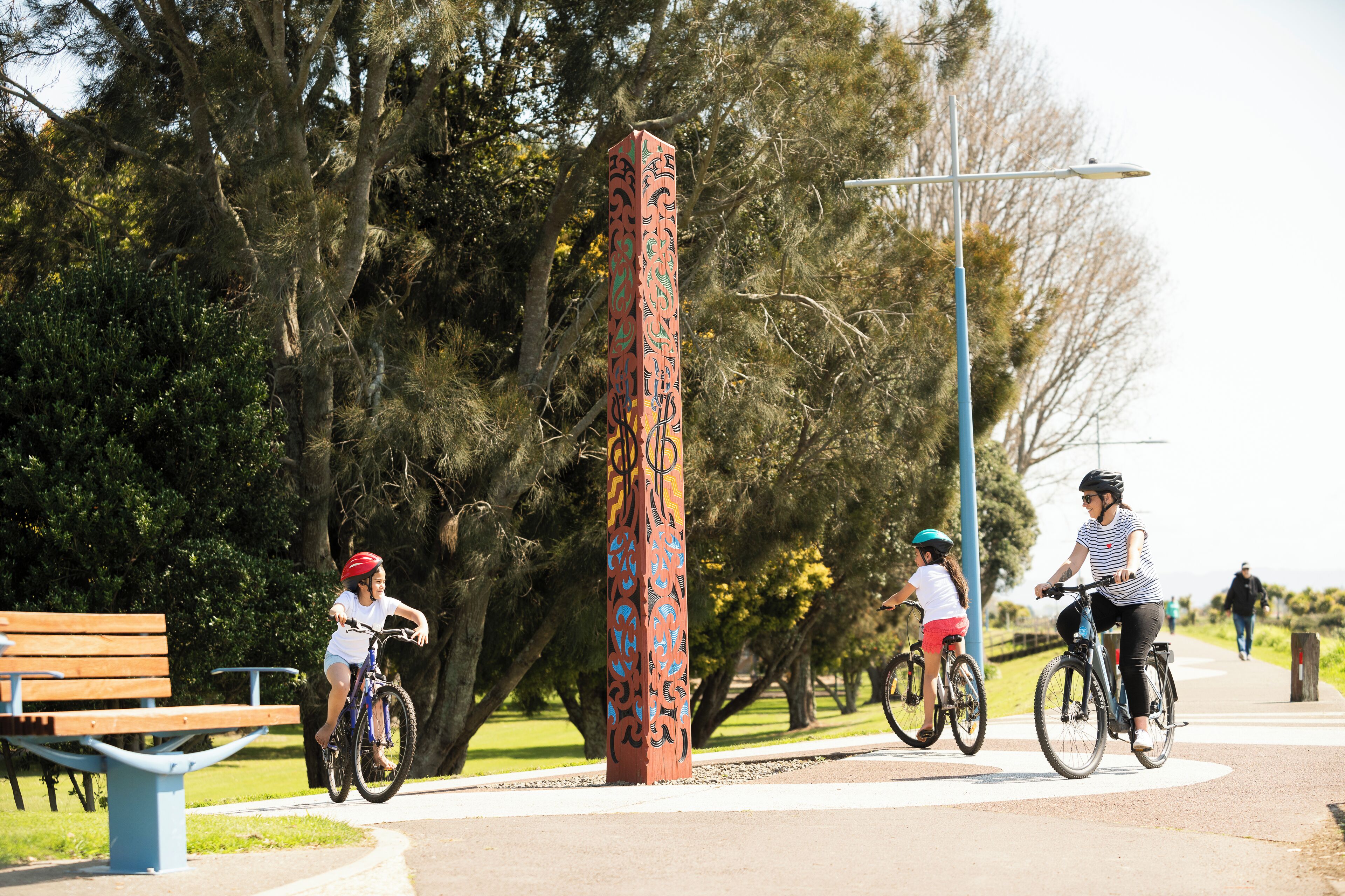 A park with a wooden bench, a tall decorative post, and cyclists on a paved path.