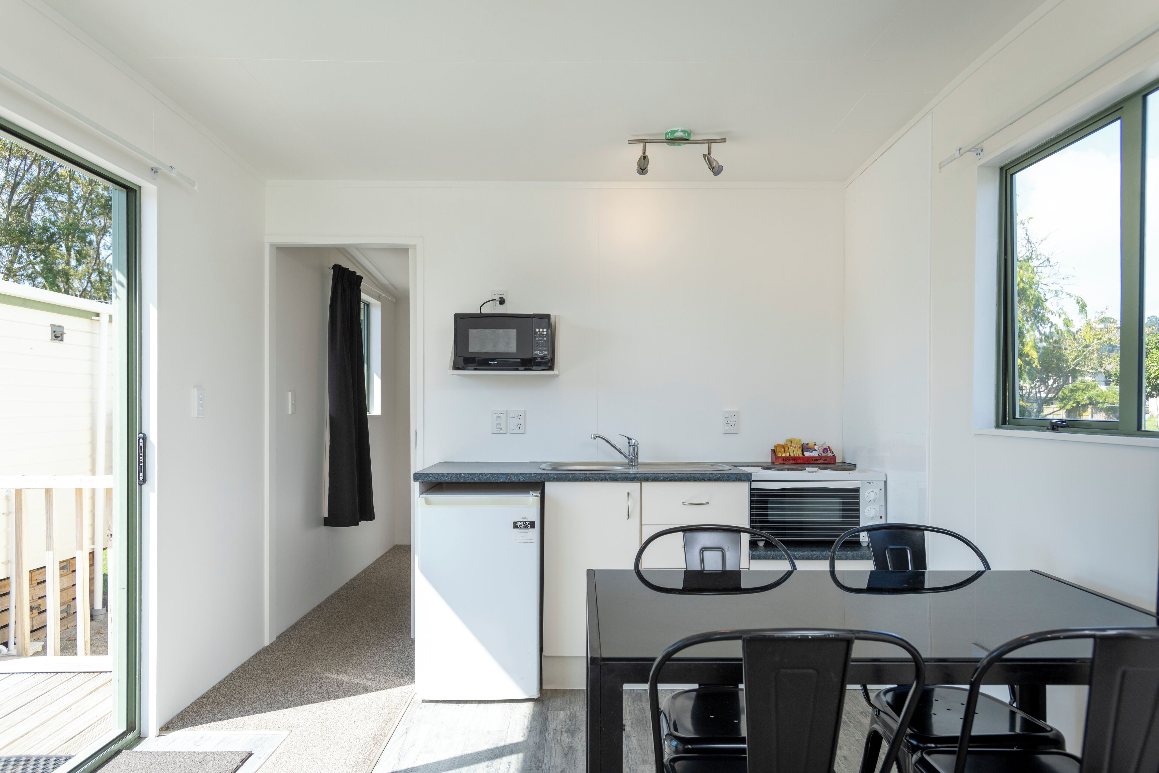 Een kleine keuken met een magnetron, spoelbak en een kleine koelkast. Er is een eettafel met stoelen en een raam dat voor natuurlijk licht zorgt.