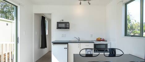A compact kitchen with a microwave, sink, and small refrigerator. There is a dining table with chairs and a window providing natural light.