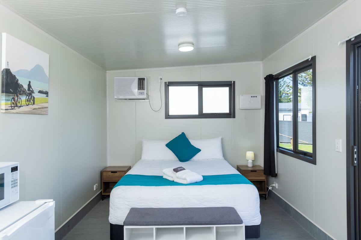 Een keurig opgemaakt hotelkamer met een bed, nachtkastjes, een wandairconditioner, een raam met gordijnen en een schilderij aan de muur.