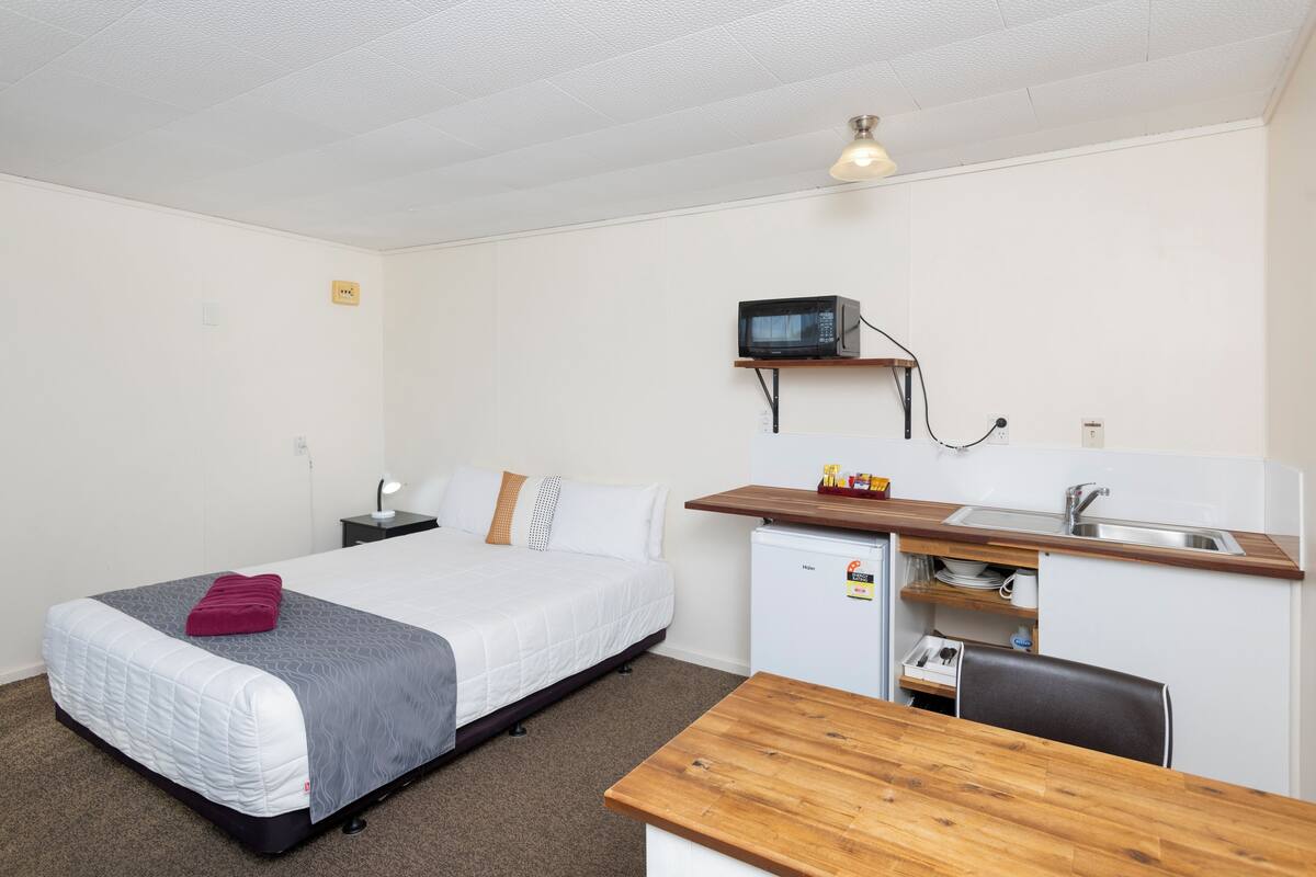 Een compacte hotelkamer met een enkel bed, een kleine kitchenette, een houten bureau en een stoel.
