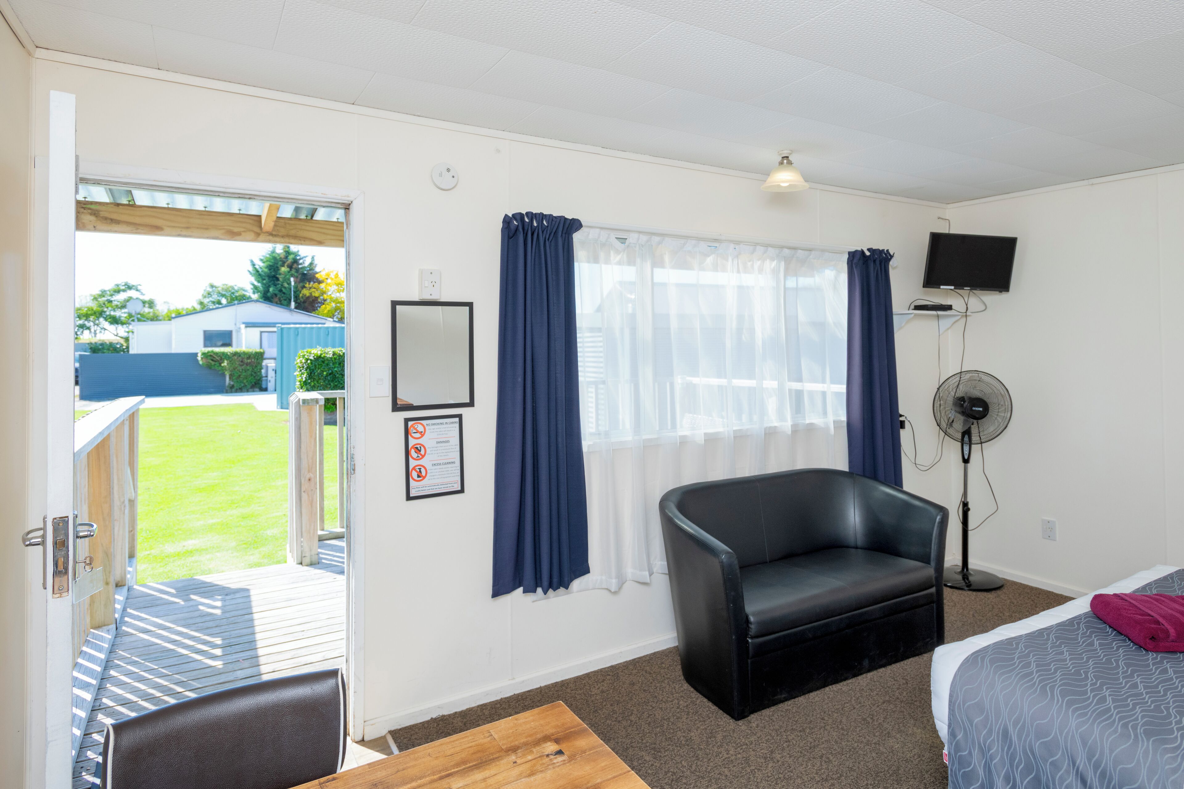 A hotel room with a bed, a black leather chair, a wooden table, a TV, a fan, and a view of the outside.