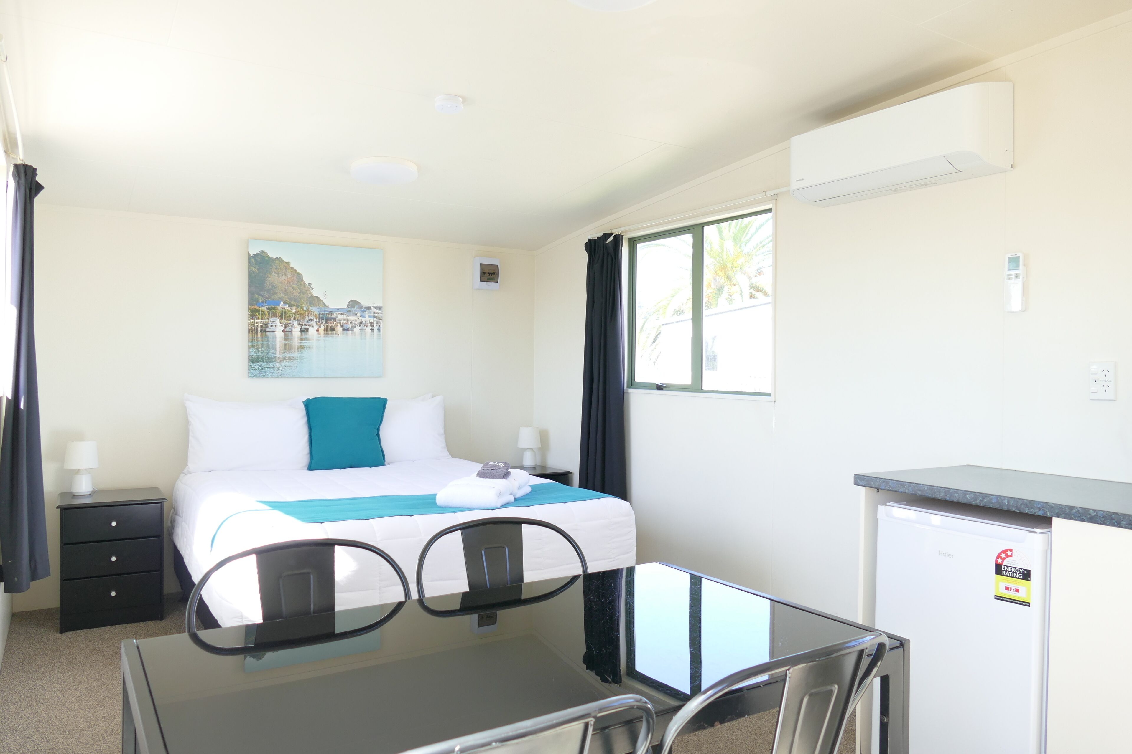 Habitación de hotel con cama, escritorio, silla, refrigerador pequeño y ventana con cortinas.