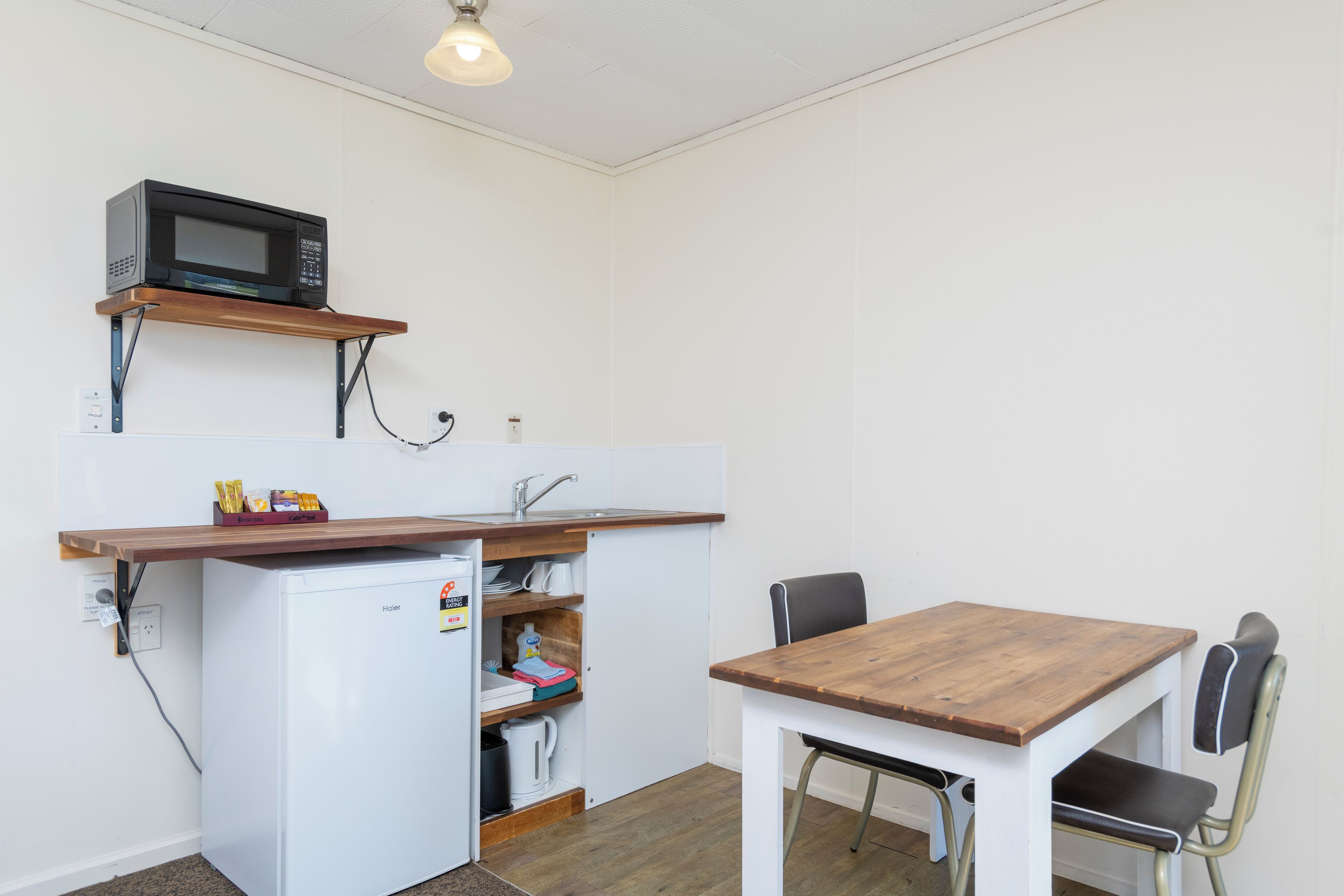 A compact kitchen with a microwave on a shelf, a refrigerator, a wooden table, and two chairs.
