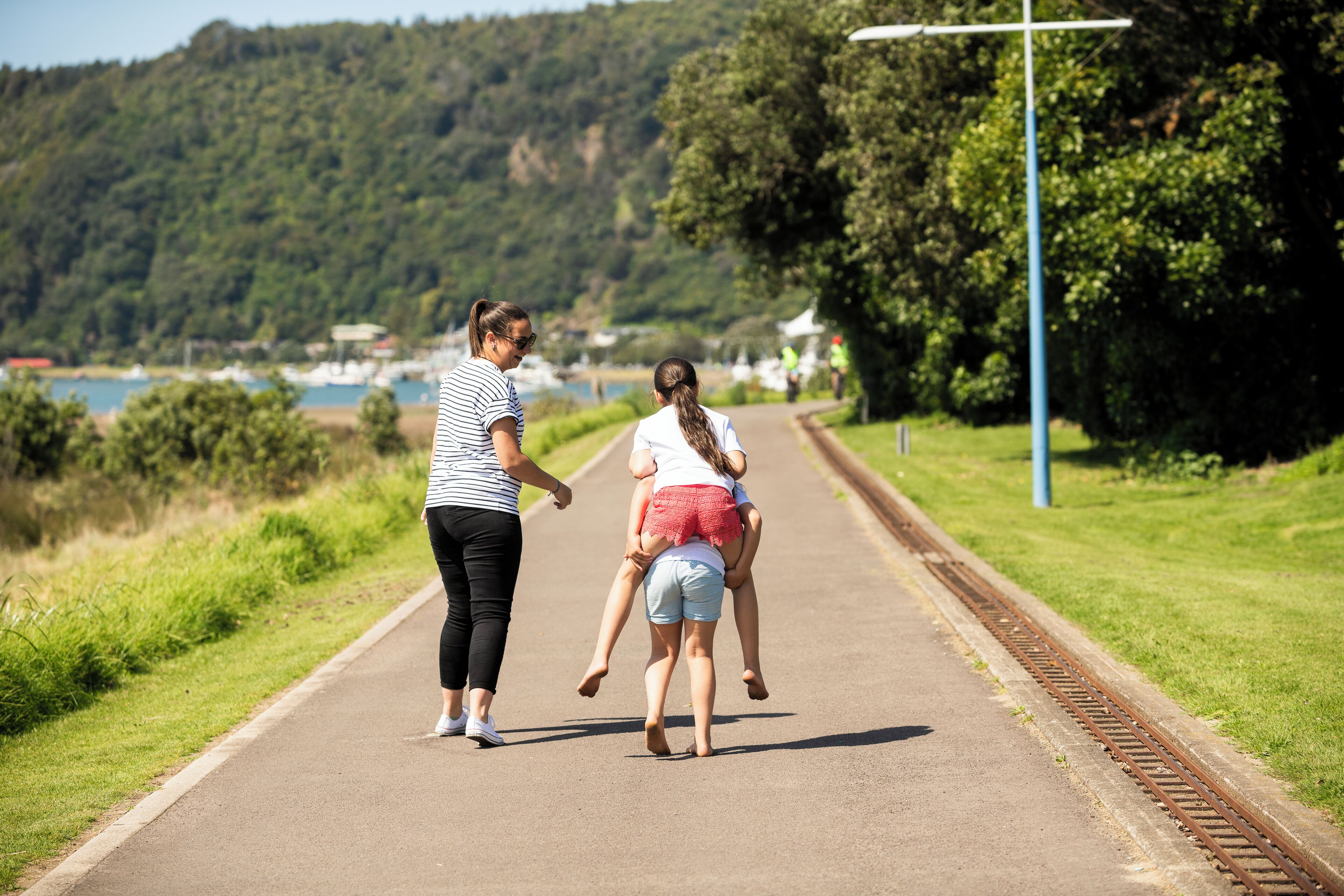 A pathway with a railway track on the side, surrounded by greenery and a body of water in the background.