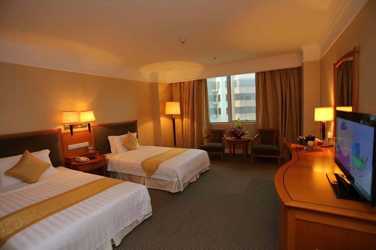 A hotel room with two beds, a desk with a TV, a small table with flowers, and a window with curtains.