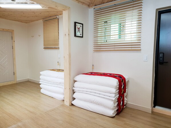 Una habitación con piso de madera, una cama con ropa blanca, una manta roja y amarilla, persianas de madera y una puerta con cerradura.
