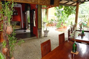 Un comedor interior con mesas y sillas de madera, rodeado de plantas en macetas y vistas al exterior.