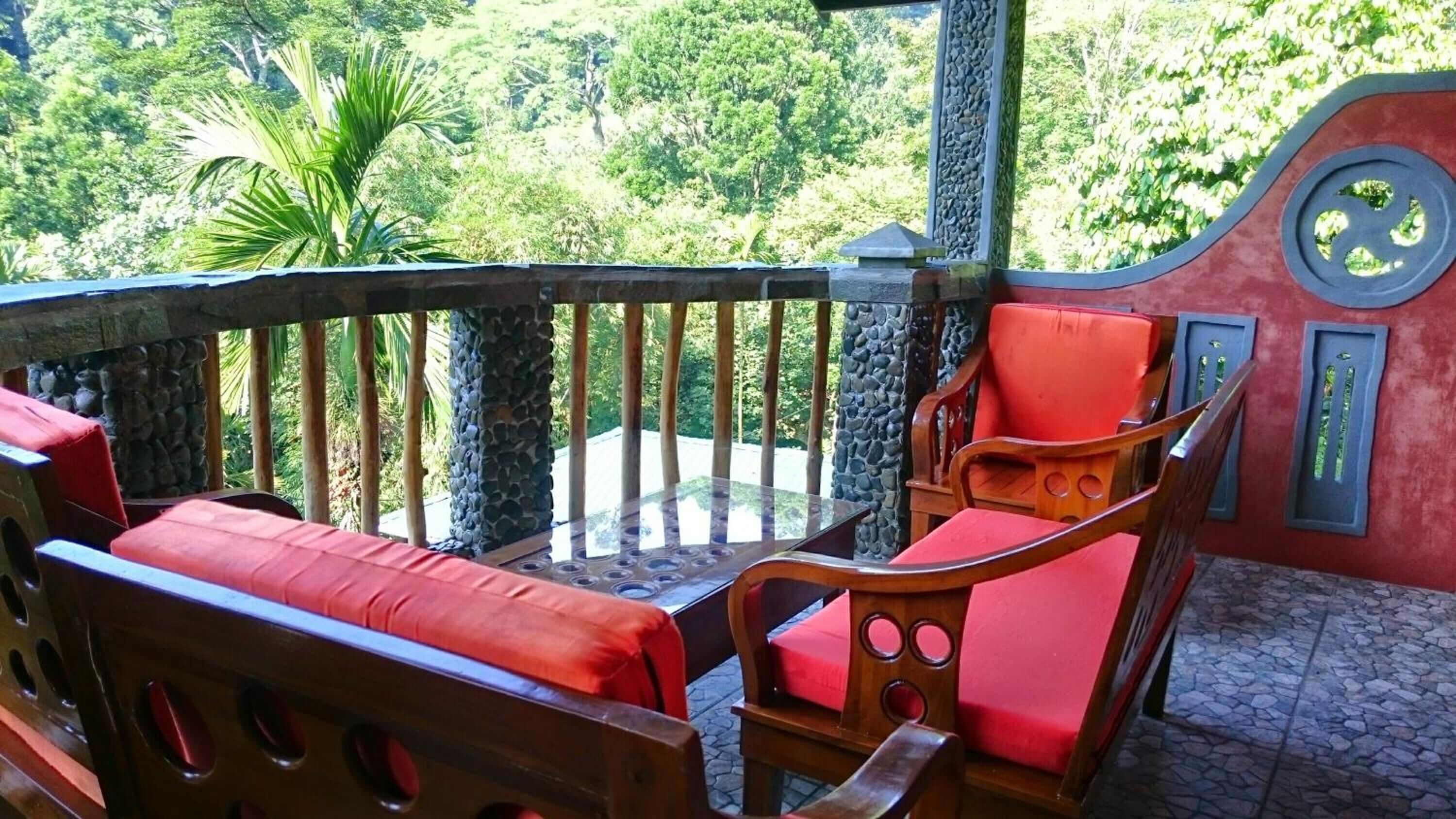 Un balcon agrémenté d’un banc en bois et d’un fauteuil rouge avec coussin, entouré d’un mur de pierre et d’une végétation luxuriante.