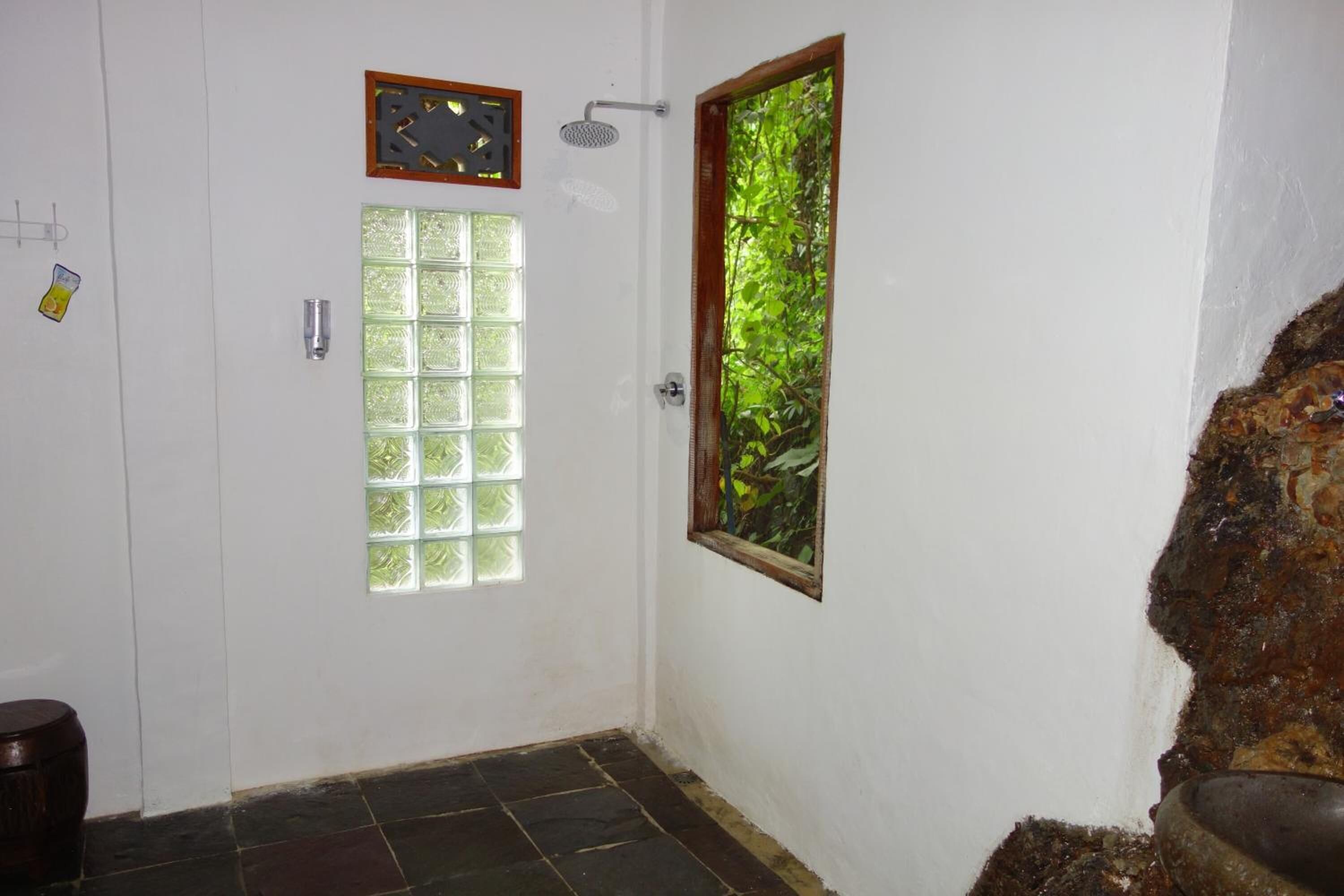 Un baño con una puerta blanca, una ventana que da a zonas verdes, una ducha y un lavabo de piedra.