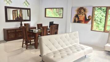 A modern living room with a white sofa, a dining table with chairs, a wooden cabinet, a large mirror, and two windows with views of greenery.