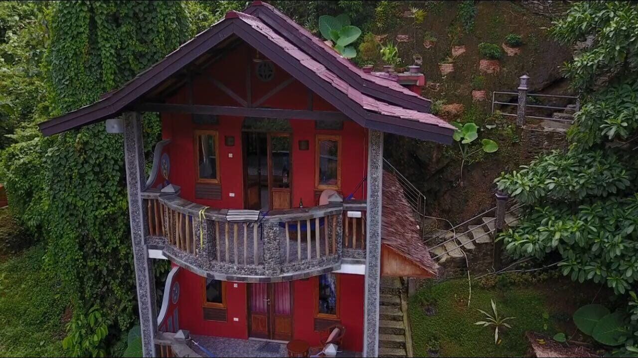 A red two-story house with a balcony, surrounded by lush greenery.