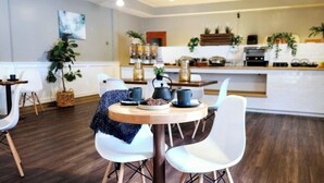 A modern dining area with a round wooden table, white chairs, and a kitchen in the background. - Western Lodge (Kimberley)