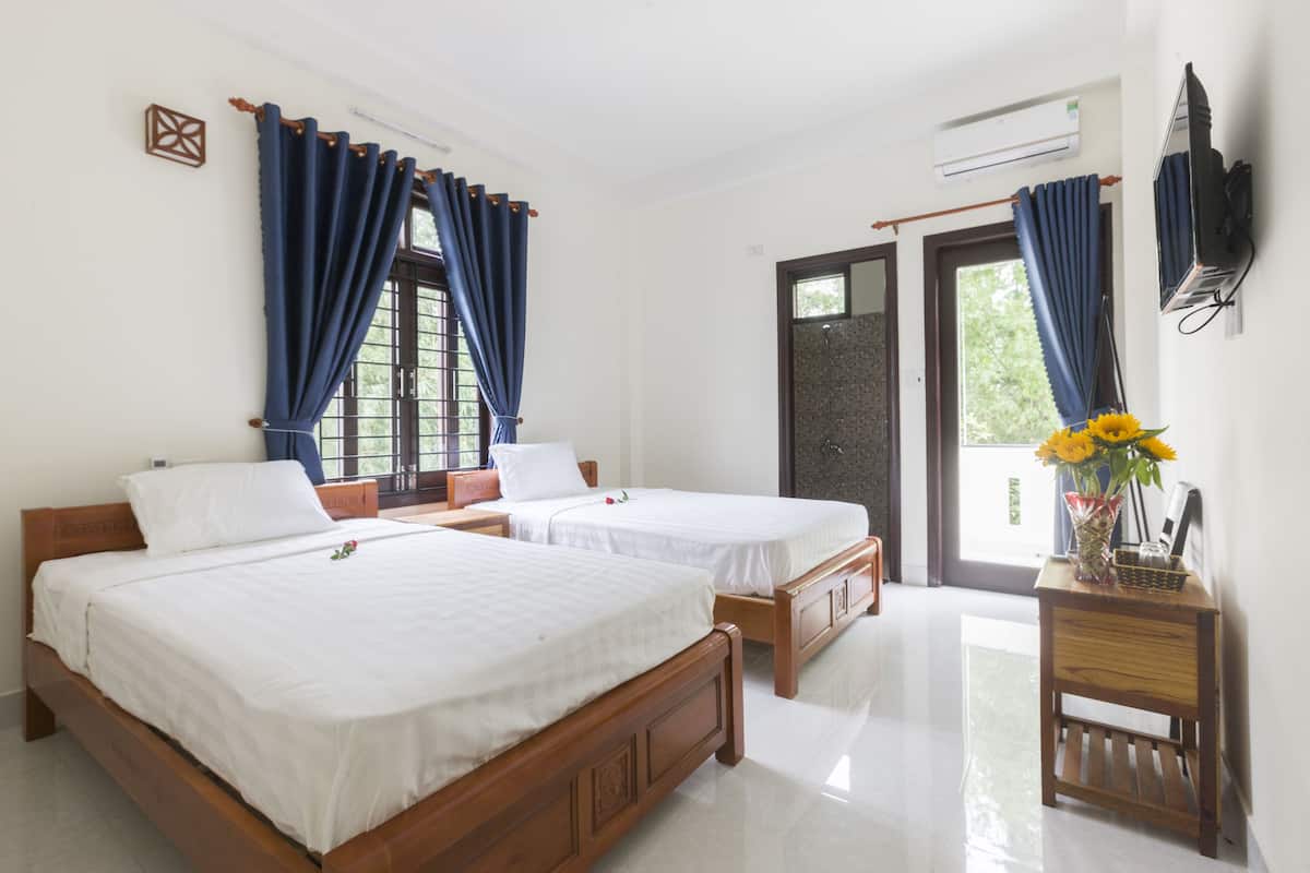 A hotel room with two wooden beds, a small wooden side table, a TV mounted on the wall, and a sliding glass door leading to an outdoor area.