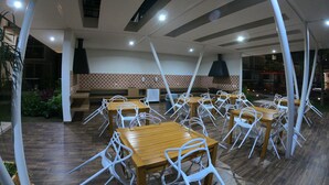 An outdoor dining area with multiple wooden tables and white chairs, surrounded by a tiled floor and a patterned wall. - Riviera Maya Apartment at Bahia Principe (Akumal)