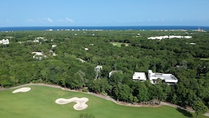 An aerial view of a golf course with a large sand bunker, surrounded by dense greenery and a few buildings. - Riviera Maya Apartment at Bahia Principe (Akumal)