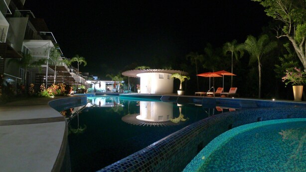A nighttime view of a swimming pool with lounge chairs and a covered area, surrounded by palm trees and a modern building. - Riviera Maya Apartment at Bahia Principe (Akumal)