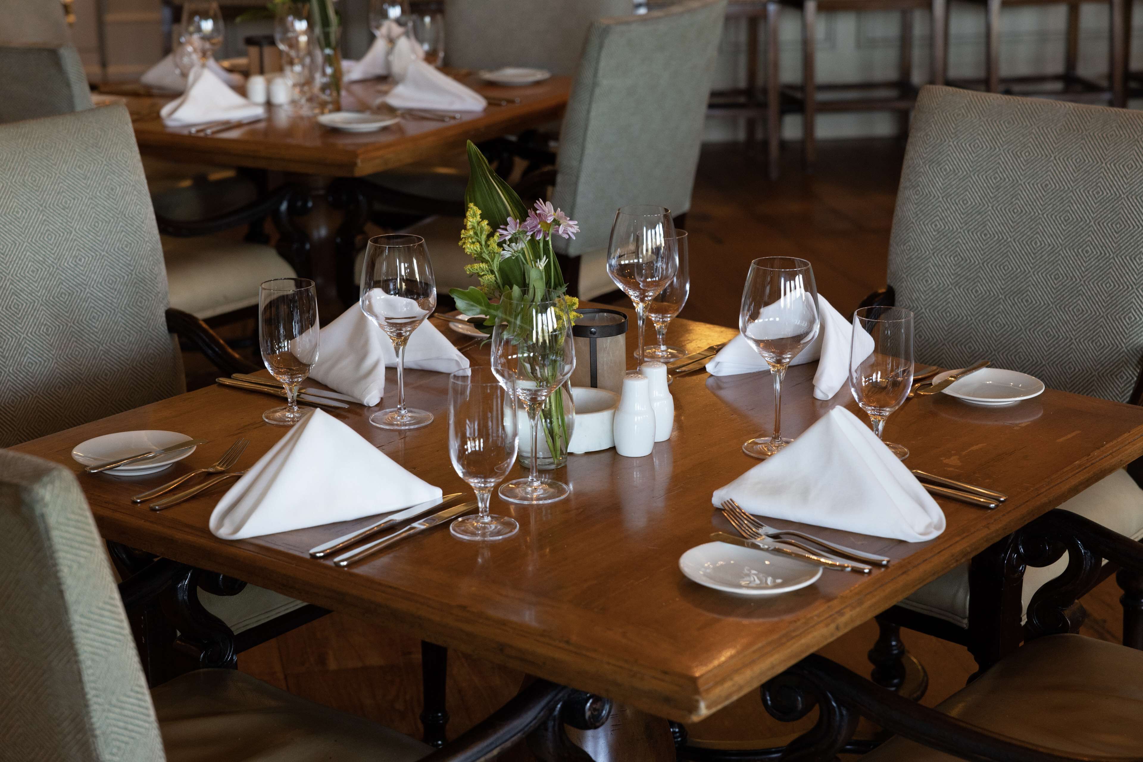 A dining table set with white napkins, silverware, and wine glasses, surrounded by upholstered chairs.