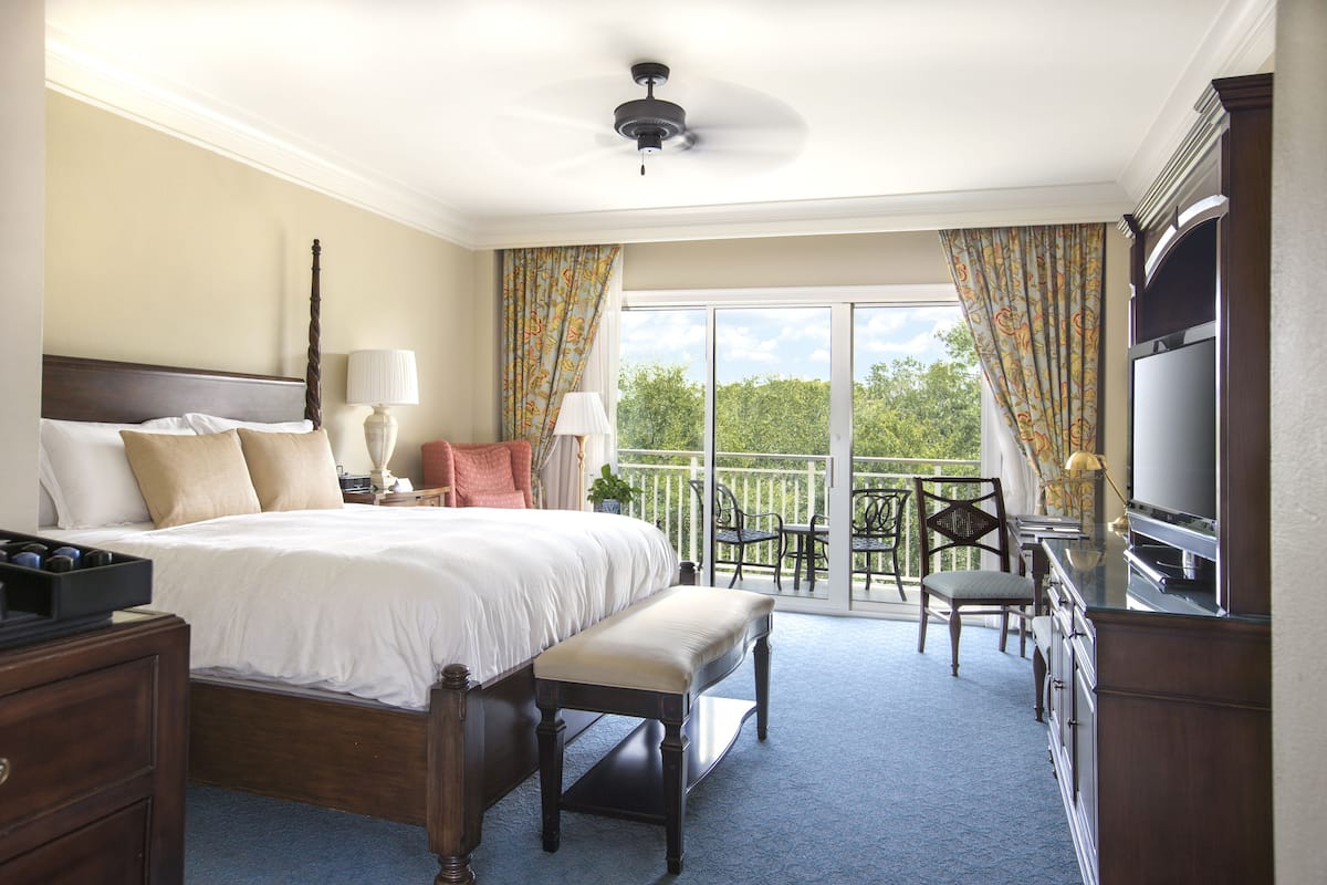 A hotel bedroom with a large bed, a bench, a TV, and a balcony with outdoor furniture.